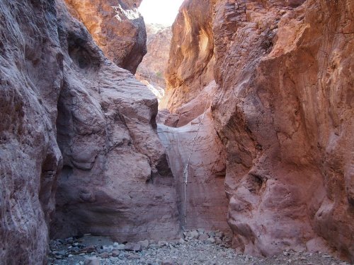 Narrow canyon with steep, reddish rock walls and a dry riverbed at the bottom, illuminated by sunlight.