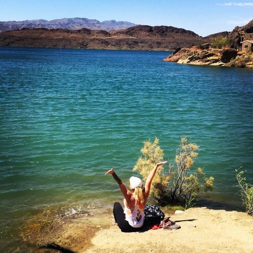 A person sits by a lake, arms raised, enjoying the scenic view of water and mountains on a sunny day.