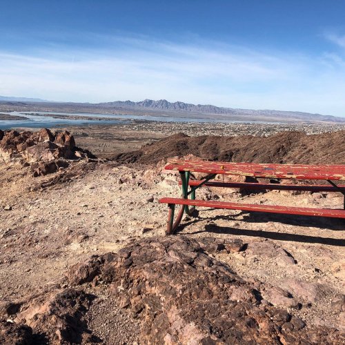 A red picnic table on rocky terrain overlooking a vast landscape with mountains and a lake in the distance.