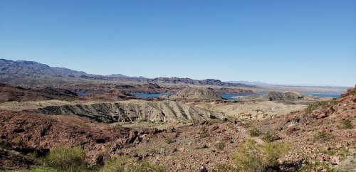 A panoramic view of rugged terrain with rolling hills and a blue lake under a clear blue sky.