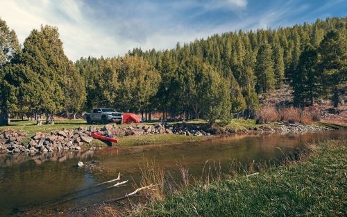 A serene campsite by a river, featuring a kayak, a tent, and a vehicle surrounded by lush trees and a clear sky.