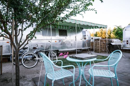 A cozy outdoor space with a silver trailer, bicycles, pink flamingos, and a table with chairs under a green awning.