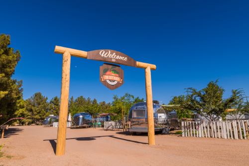 A wooden sign reading "Welcome" stands at the entrance of a campground with Airstream trailers and blue skies.