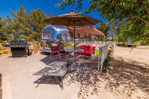 A shiny airstream trailer under an umbrella, with a table, chairs, and a grill in a sunny outdoor setting.