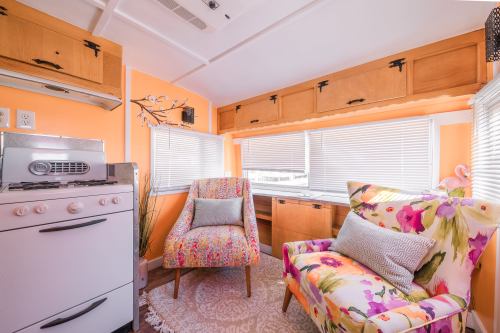 Cozy interior of a colorful trailer with orange walls, floral chairs, a small kitchen area, and bright natural light.