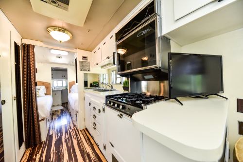 Interior of a modern camper kitchen, featuring a stove, microwave, and a small TV, with a view of the sleeping area.