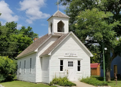 A small white historic schoolhouse with a bell tower, surrounded by greenery and a clear blue sky.