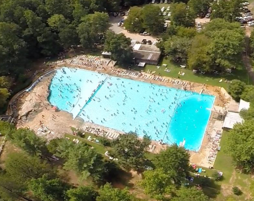 Aerial view of a large swimming pool surrounded by trees, with people swimming and lounging on the deck.