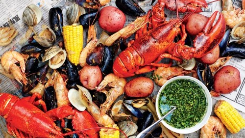 A seafood feast featuring lobster, shrimp, mussels, clams, corn, and potatoes on newspaper, with a bowl of green sauce.