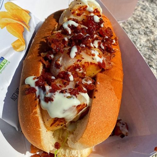 A hot dog bun filled with grilled scallops, topped with bacon bits and a creamy sauce, served with potato chips.