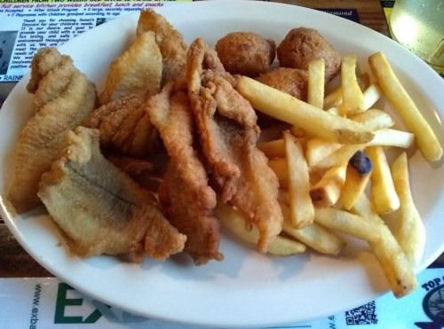 A plate of fried fish fillets, golden French fries, and hushpuppies served on a white dish.