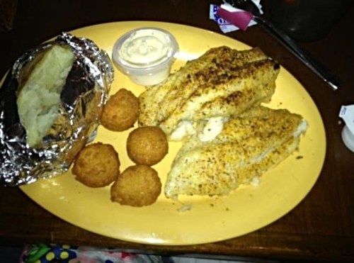 A plate with two seasoned fish fillets, a baked potato, cornbread balls, and a small cup of dipping sauce.