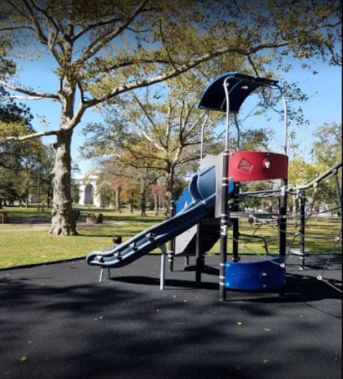 A playground slide and climbing structure in a park, surrounded by trees and a clear blue sky.
