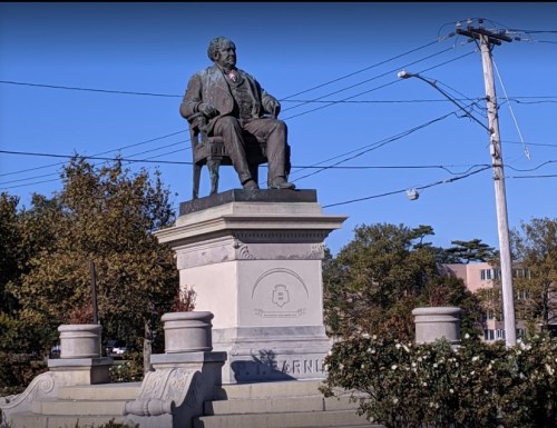 A bronze statue of a seated man on a pedestal, surrounded by trees and power lines.