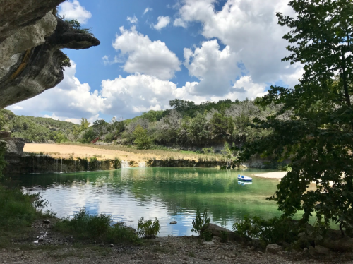 A serene lake surrounded by greenery and trees, with a small boat on the water under a partly cloudy sky.