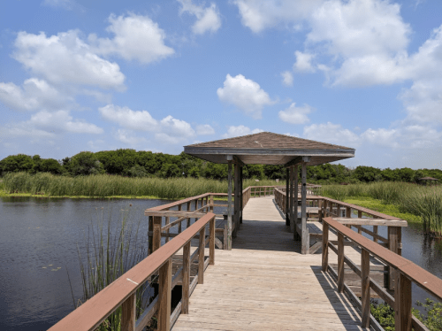 A wooden boardwalk leads to a gazebo over a calm waterway, surrounded by lush greenery and blue skies.