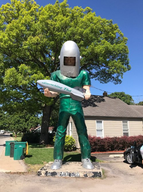 A large statue of a man in a green suit holding a model rocket, standing in front of a house and tree.