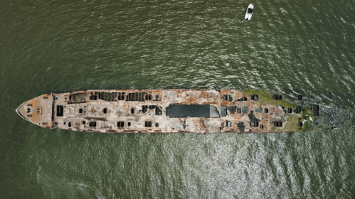 Aerial view of a rusted shipwreck partially submerged in green water, with a small boat nearby.