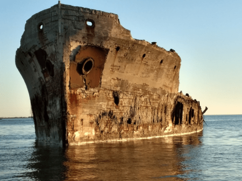 A weathered shipwreck partially submerged in calm waters, with rusted metal and a clear sky in the background.