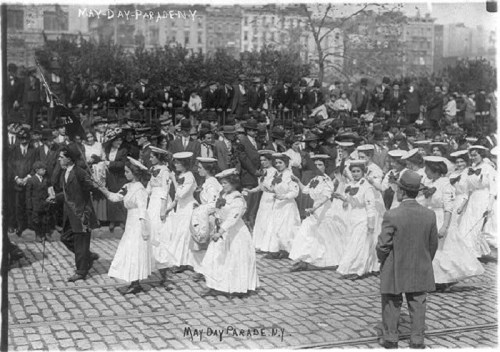 Black and white photo of a May Day parade, featuring women in white dresses and a crowd of onlookers in hats.