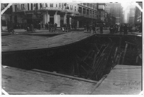 A historic black-and-white photo of a street with a large, collapsed area revealing wooden structures beneath.