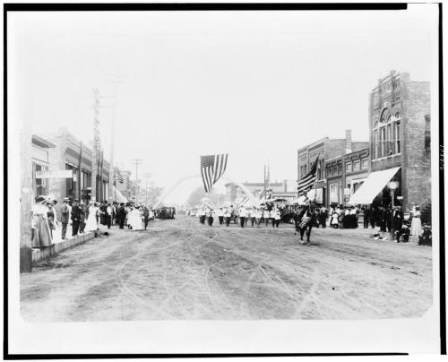 A historic parade scene with people lining the street, an American flag hanging overhead, and buildings in the background.