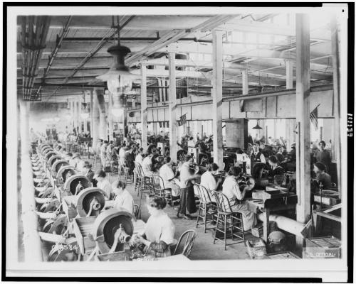 A historic black-and-white photo of a factory interior with workers at sewing machines, focused on their tasks.