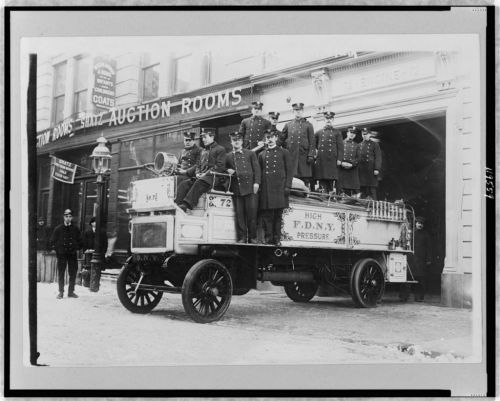Historic black-and-white photo of firefighters on a vintage fire truck outside an auction house.