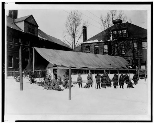 A black and white photo of a group in winter clothing, standing in the snow near a large tent and buildings.