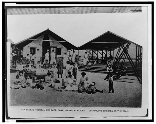 Black and white photo of Sea Breeze Hospital, Coney Island, with children and adults on the beach in front of the buildings.
