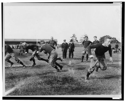 Historic black and white photo of young football players practicing on a grassy field, with spectators in the background.