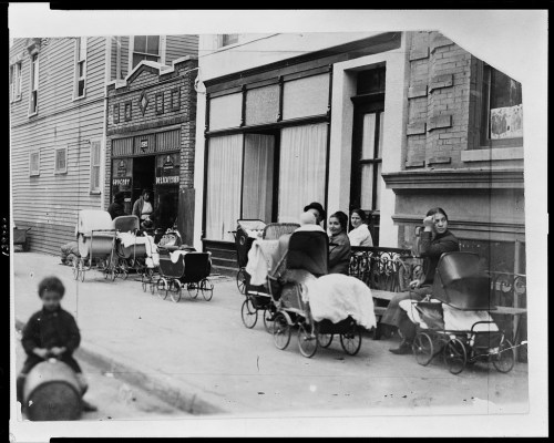 A historic black-and-white photo of women with baby carriages on a city street, with children playing nearby.