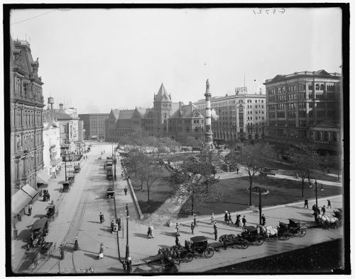 Historic city square with trees, buildings, and people walking, featuring horse-drawn carriages and a statue in the background.