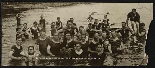 A vintage photo of a group of people in swimsuits enjoying a lake, some standing in water, others on the shore.