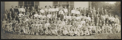 A large group photo of children and adults, gathered outside a building, in early 20th-century attire.