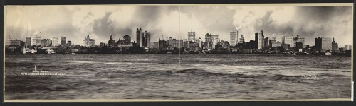 Panoramic view of a city skyline with tall buildings and boats on the water, under a cloudy sky.