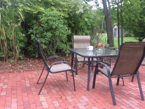 A patio with a glass table, two chairs, a coffee cup, and flowers, surrounded by greenery and brick flooring.
