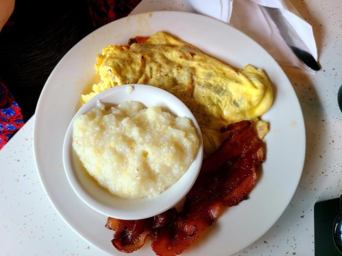 A plate with a fluffy omelet, crispy bacon, and a bowl of creamy grits.
