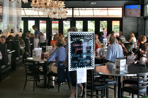 A busy restaurant interior with patrons dining, a chalkboard sign, and a chandelier overhead.