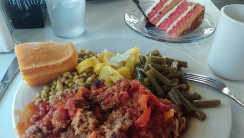 A plate of meatloaf with tomato sauce, green beans, squash, peas, cornbread, and a slice of pink cake on the side.