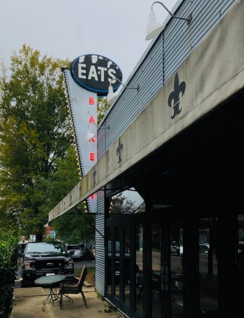A restaurant exterior with a sign reading "EATS," featuring outdoor seating and a tree in the background.