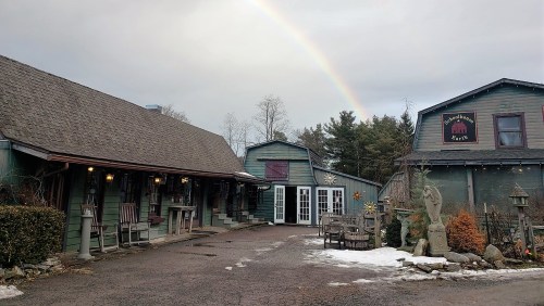 A rustic building with a porch, surrounded by trees, under a cloudy sky with a rainbow in the background.