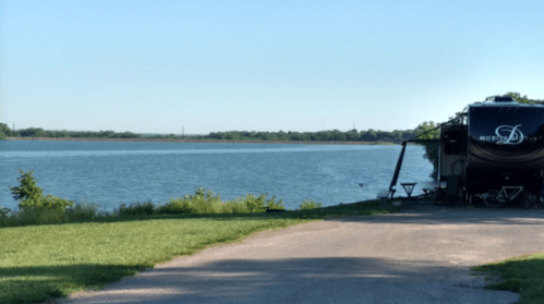 A serene lakeside view with an RV parked on a grassy area, surrounded by trees and clear blue skies.