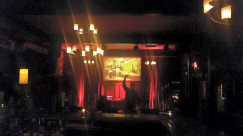 A dimly lit bar with red lighting, featuring a DJ booth and a large screen displaying a vibrant image.