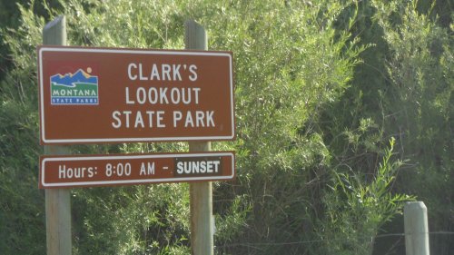 Sign for Clark's Lookout State Park in Montana, displaying park hours from 8:00 AM to sunset. Surrounded by greenery.