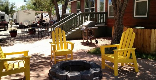 A cozy outdoor space with three yellow chairs around a stone fire pit, near a wooden house and picnic area.
