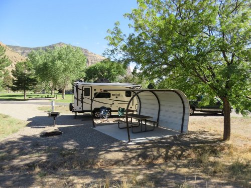 An RV parked near a shaded picnic area with a table and grill, surrounded by trees and mountains in the background.
