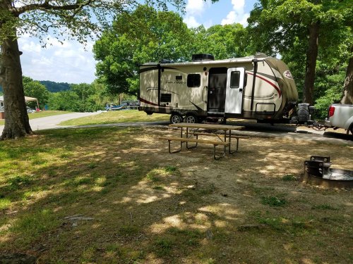 A parked RV near a picnic table and fire pit, surrounded by trees and a scenic landscape.