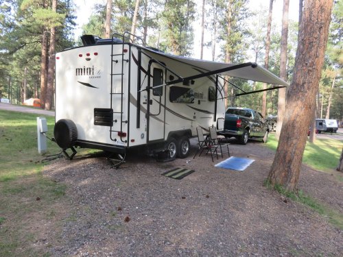 A small camper trailer parked under trees, with an awning extended and outdoor seating set up nearby.