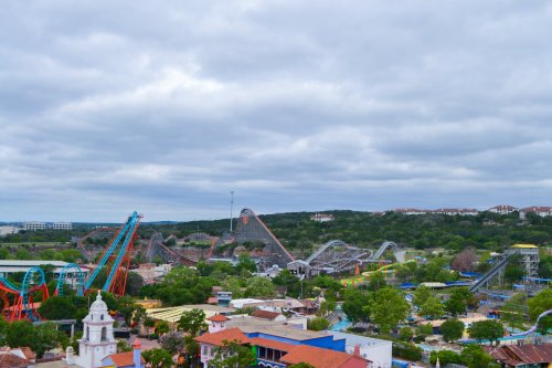 Aerial view of a theme park featuring roller coasters, green trees, and a cloudy sky in the background.